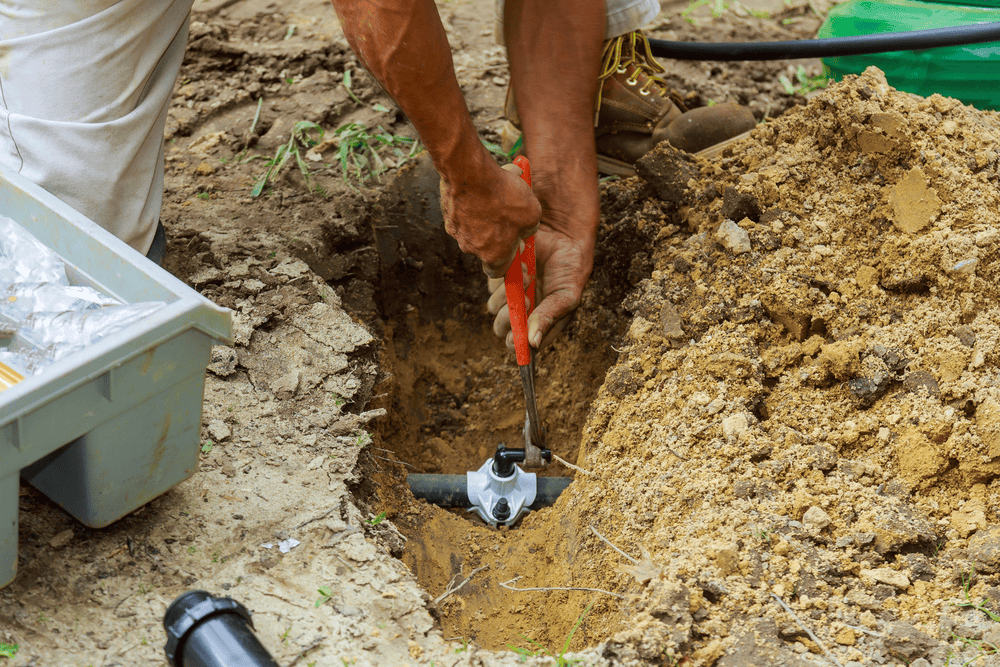 Person using a wrench to install or repair an irrigation valve in a shallow trench, surrounded by dirt and tools, with boots and plastic parts box visible nearby.