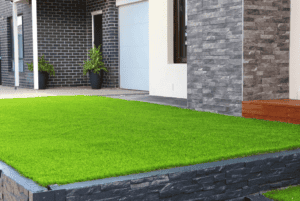 A modern house exterior with dark brick and stone walls, a white garage door, potted plants, wooden steps, and a neatly maintained lawn with bright green artificial grass in the front yard.