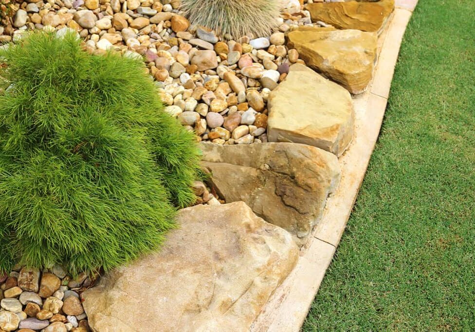 A garden bed with large rocks and pebbles borders a section of green grass. A lush, bushy green plant is surrounded by stones and gravel, creating a neat, low-maintenance landscape design.