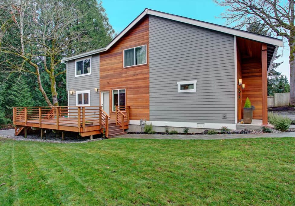 Modern two-story house with gray and wood panel siding, large windows, a wooden deck, and a manicured lawn designed by a Kenmore Landscaper, surrounded by trees under a clear sky.