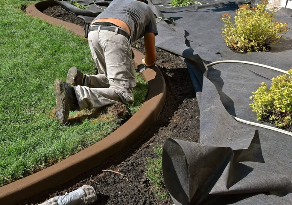 A person kneels on the grass while installing a curved concrete landscape edging between a lawn and a garden bed covered with black landscaping fabric—exemplifying quality work by an Issaquah Landscaping Company. Shrubs and a coiled white hose are nearby.