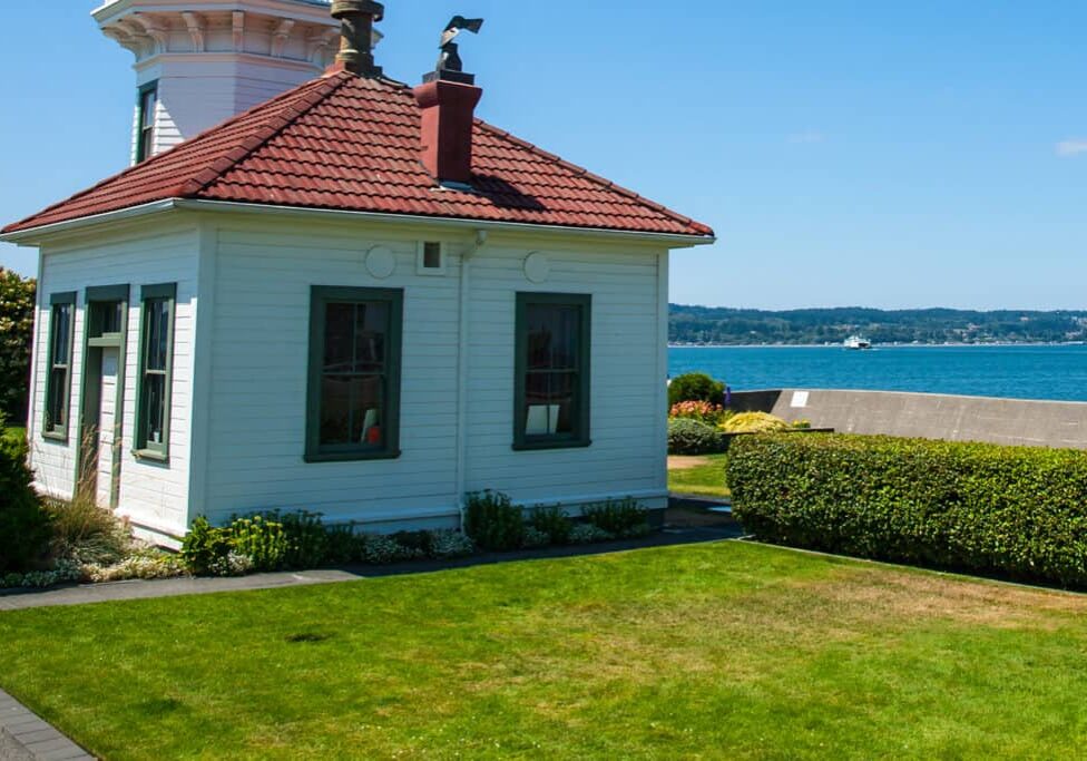 A small white building with green-trimmed windows and a red-tiled roof sits by neatly trimmed green grass, overlooking a blue body of water under a clear sky.