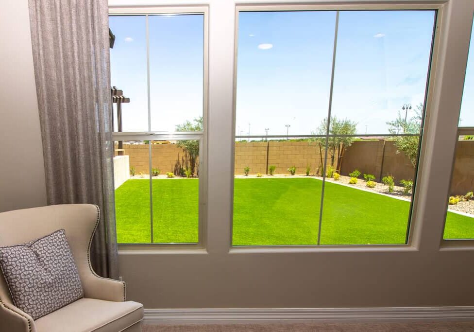A bright room with a beige armchair and patterned pillow sits by large windows, showcasing a green, neatly landscaped backyard by Queen Anne Landscaping Company, with a brick wall and small trees under a clear blue sky.